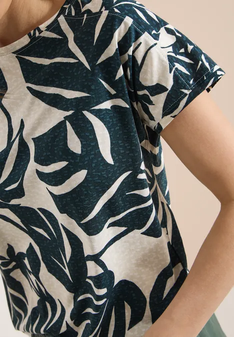 Nahaufnahme eines beigen T-Shirts mit dunkelblauem, abstraktem Blattmuster auf Schulter und Arm.