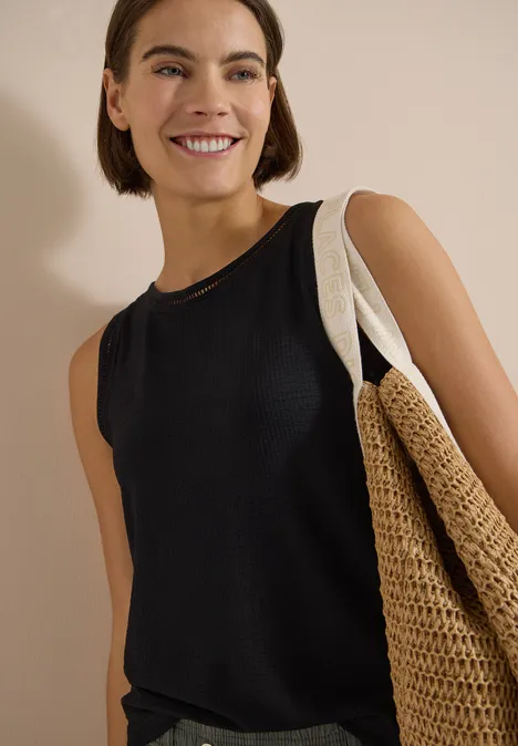 Top mit U-Boot-Ausschnitt und Spitzendetails Black Frau in schwarzem ärmellosem Top mit großem geflochtenem Strohtasche vor neutralem Hintergrund.
