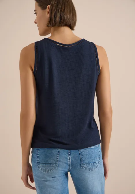 Frau von hinten in ärmellosem dunkelblauem Top und blauen Jeans vor neutralem Hintergrund.