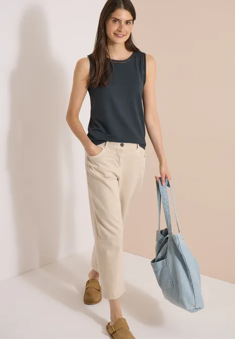 Top mit U-Boot-Ausschnitt und Spitzendetails palm breeze green Frau trägt dunkles ärmelloses Top, helle Hose und blaue Stofftasche.