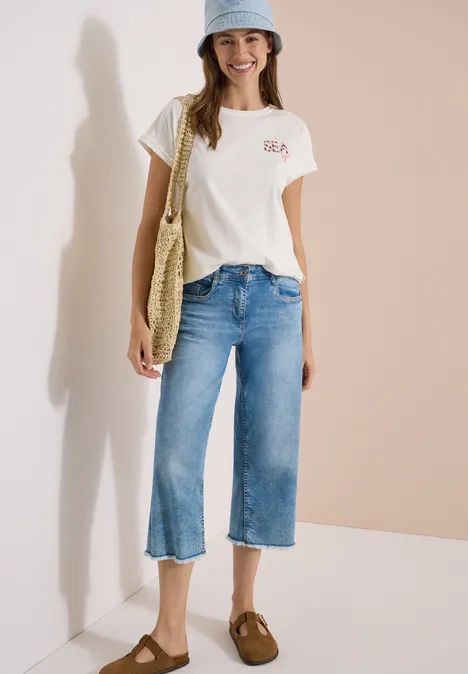 T-Shirt mit Rundhals und Embroidery vanilla white Frau in weißem T-Shirt, weiten Jeans-Culottes, Basecap, braunen Slippern und Strohtasche vor beigem Hintergrund.