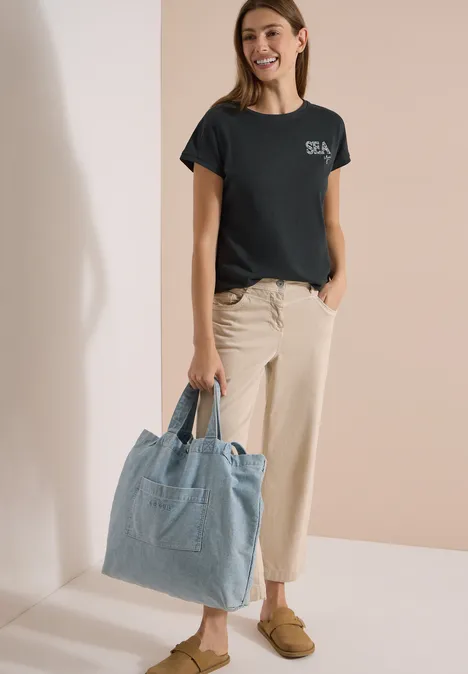 T-Shirt mit Rundhals und Embroidery palm breeze green Frau trägt schwarzes T-Shirt, beige Hose, braune Loafers und hält eine hellblaue Jeans-Tasche.