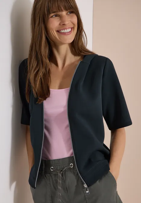 Frau lehnt an Wand, trägt schwarzen Cardigan über rosa Top und graue Hose.