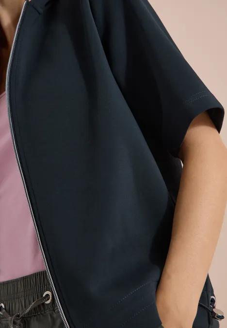 Person trägt eine dunkelblaue, kurzärmlige Jacke über einem rosa Top und lockere, graue Bundhose.