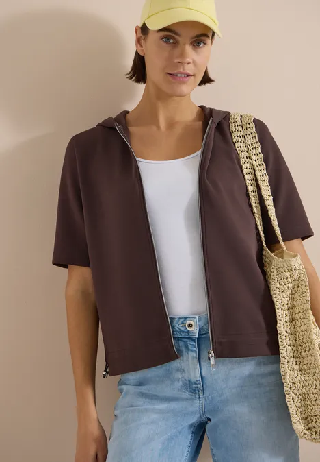 Kapuzenjacke mit seitlcihen Tunnelzügen brown cacao Person trägt kurzes braunes Kapuzenshirt, weißes Top, blaue Jeans und Basttasche.