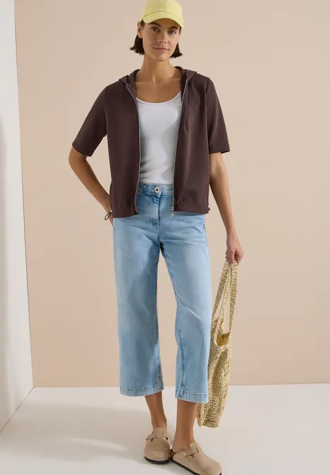 Kapuzenjacke mit seitlcihen Tunnelzügen brown cacao Frau mit weißem Top, kurzarm Hemd, weitem Jeans, Sandalen und Basttasche in minimalistischem setting.
