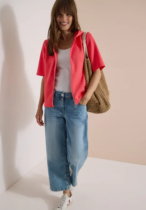 Frau trägt weißes Top, pinke Jacke, weite Jeans, weiße Sneaker und Korbtasche vor hellen Wänden.