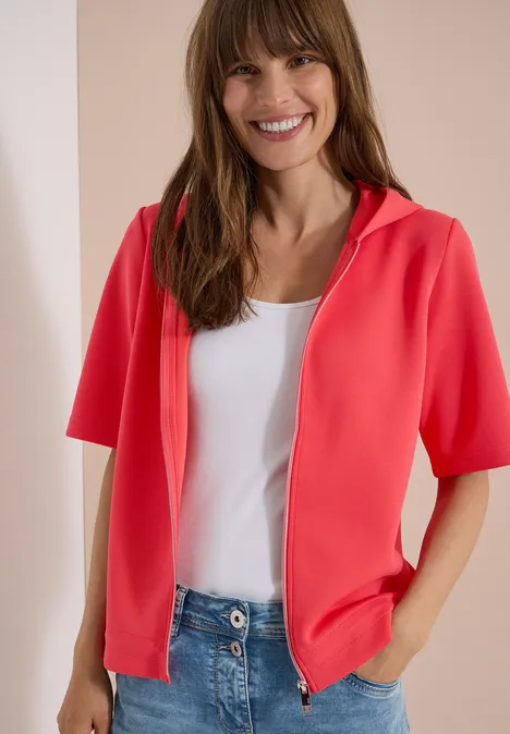 Person trägt korallenrote kurzärmelige Kapuzenjacke, weißes Top und Jeans.