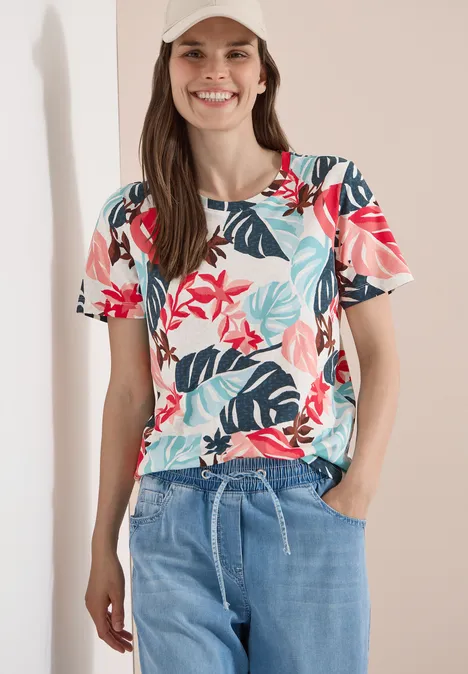 Frau trägt kurzärmlige Bluse mit rotem Blumen- und blauem Blattmuster und hellblauen Jeans.
