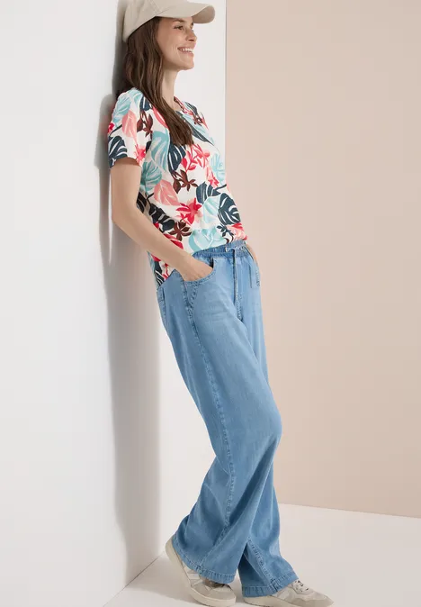Eine Person lehnt entspannt an einer Wand, trägt ein buntes Blumen-T-Shirt, weite Jeans und weiße Sneaker.