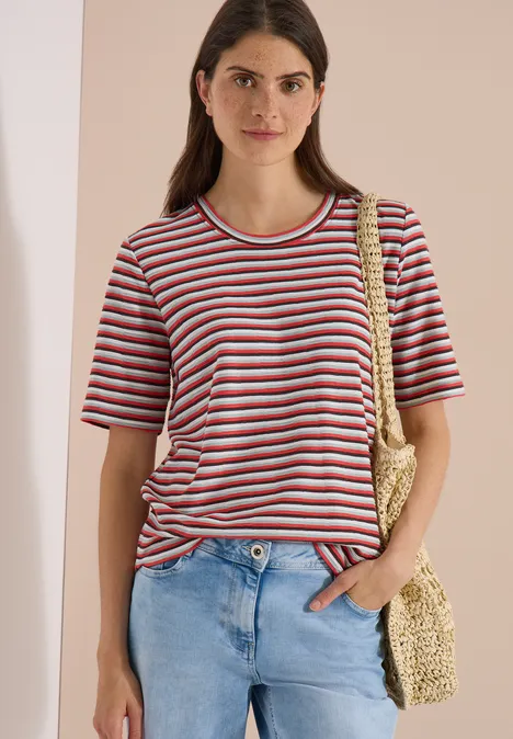 Frau in gestreiftem T-Shirt, Jeans und Strohtasche vor beigem Hintergrund.