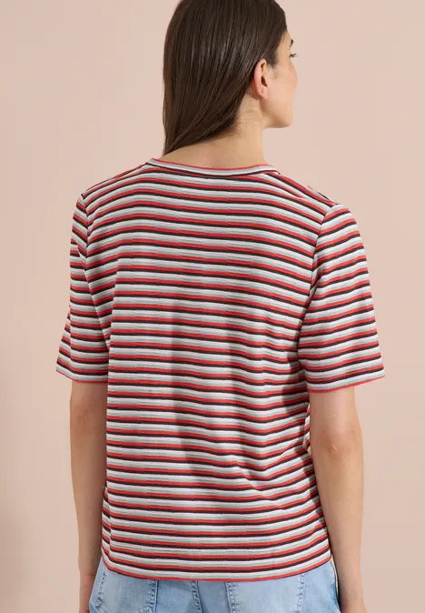 Frau von hinten, trägt gestreiftes T-Shirt mit roten, weißen und blauen Streifen vor beigem Hintergrund.