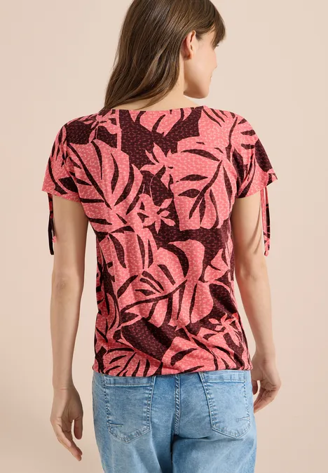 Frau von hinten in rosa-schwarzem Blusenshirt mit Blattmuster und blauen Jeans.
