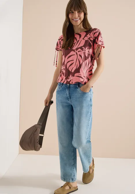 Frau trägt pinkes Blusenshirt mit tropischem Muster, helle Jeans, Bast-Sandaletten und eine Tasche.