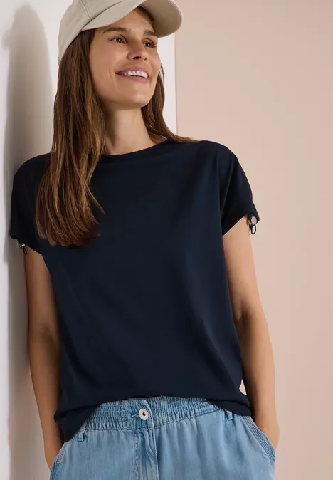 Eine Person trägt ein dunkelblaues T-Shirt, helle Jeans und eine helle Cap, lehnt an einer hellen Wand.