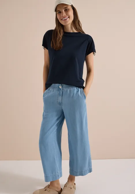 Frau in dunkelblauem T-Shirt, hellblauer Culotte-Hose und Sandalen vor neutralem Hintergrund.