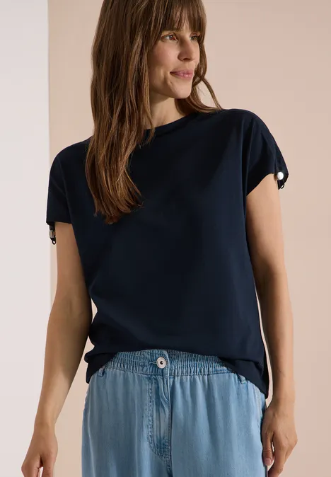 Frau in dunkelblauem T-Shirt und hellblauem Jeansrock vor beigem Hintergrund.