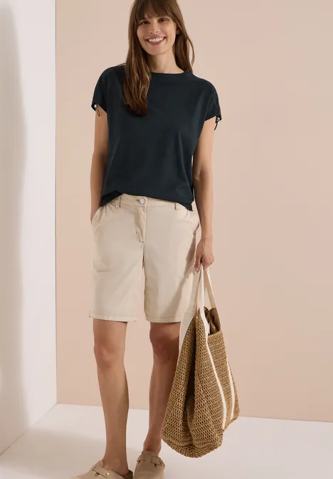 Frau trägt dunkelblaues T-Shirt, beige Bermuda-Shorts und eine Strohtasche.