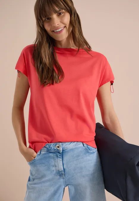 Shirt met rimpels in effen kleur coral Vrouw draagt rood T-shirt, blauwe spijkerbroek en houdt een zwarte jas vast.