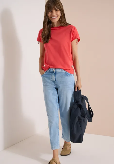Shirt met rimpels in effen kleur coral Een persoon staat voor een witte achtergrond, draagt een rood T-shirt, lichte spijkerbroek en houdt een donkere tas vast.