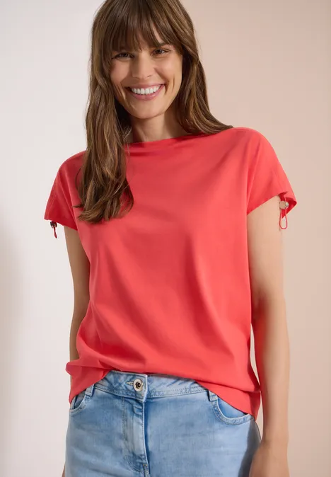 Shirt met rimpels in effen kleur coral Vrouw in een koraalroze t-shirt met korte mouwen en een spijkerbroek voor een lichte achtergrond.