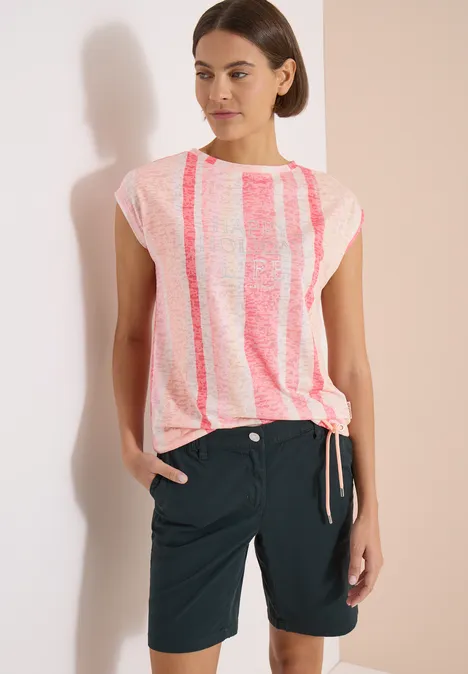 Femme en haut court rose et blanc à rayures et short foncé, posant devant un mur blanc.