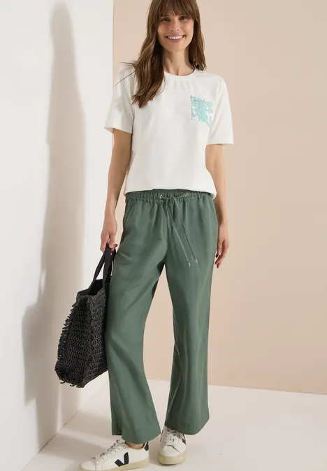 T-Shirt mit Rundhals und Brusttasche vanilla white Frau trägt weißes T-Shirt mit gemusterter Tasche, weite grüne Hose, weiße Sneaker und schwarzen Netzbeutel.