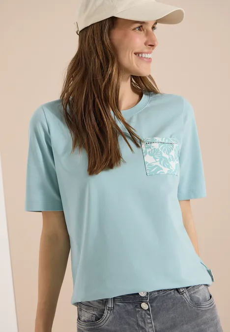 T-Shirt mit Rundhals und Brusttasche crystal mint Frau in hellblauem T-Shirt mit Brusttasche und Print, dazu Jeans und Cap vor neutralem Hintergrund.