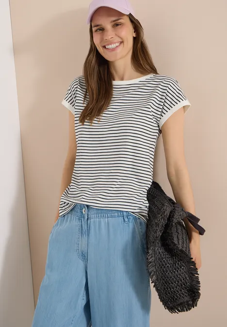 Junge Frau trägt gestreiftes T-Shirt und lockere Jeanshose, hält eine schwarze Tasche.