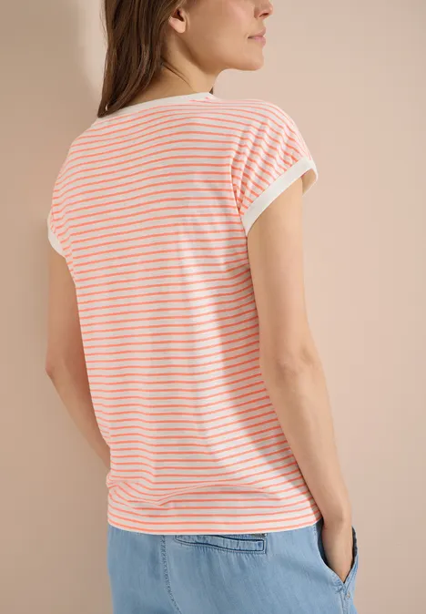 Vrouw van achteren in een wit-oranje gestreept shirt en blauwe broek tegen neutrale achtergrond.