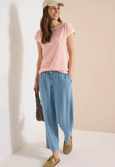 Vrouw in roze T-shirt, losse lichtblauwe broek en instappers, voor een beige muur.