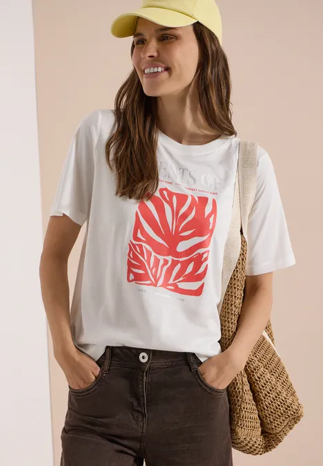 Shirt met ronde hals en print op de voorkant vanilla white Vrouw met gele pet, wit shirt met rood bladerenmotief, bruine broek en rieten tas voor een lichte achtergrond.