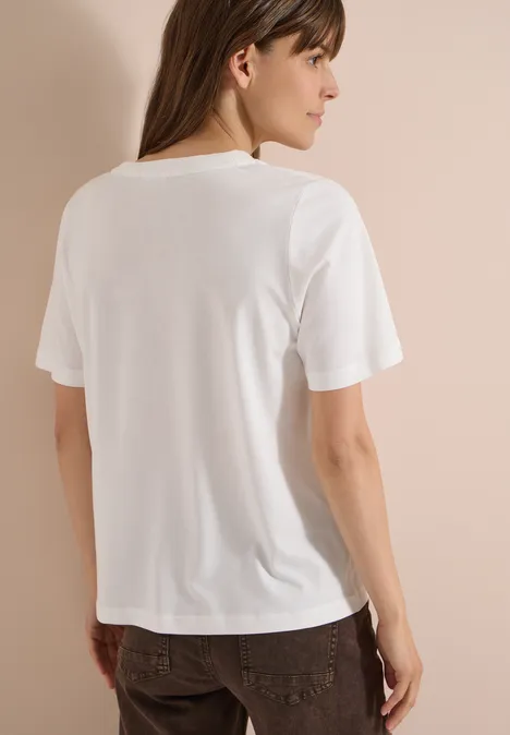 Shirt met ronde hals en print op de voorkant vanilla white Vrouw met rug naar de camera in een wit T-shirt en bruine broek voor een beige achtergrond.