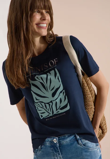 Vrouw draagt donkerblauw t-shirt met groen bladprint, spijkerbroek en gevlochten schoudertas.
