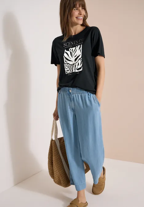 T-Shirt mit Rundhals und Frontprint palm breeze green Frau in schwarzem T-Shirt mit Zebramuster, blauen weiten Hosen und Basttasche