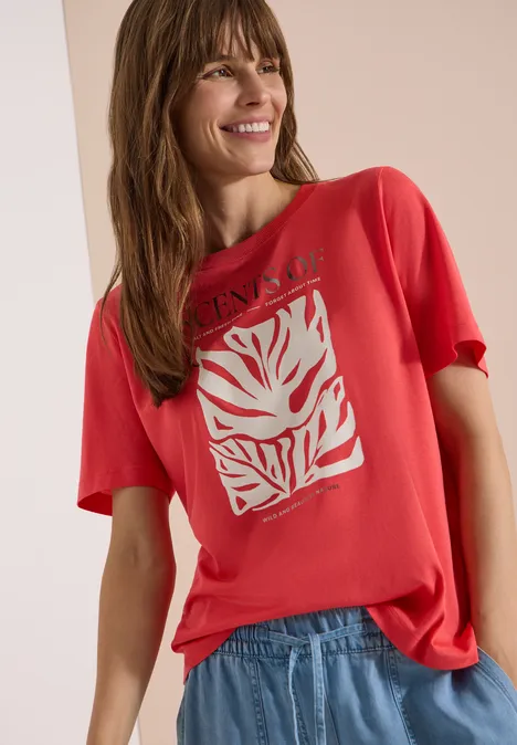 T-Shirt mit Rundhals und Frontprint coral Frau mit rotem T-Shirt mit weißem grafischen Motiv und blauer Jeans, stehend vor neutralem Hintergrund.