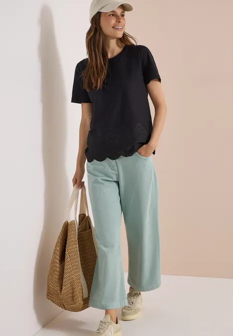 Frau in schwarzem kurzärmligem Shirt mit gewelltem Saum, mintfarbener weiter Hose, Strohtasche, Sneakern und Cap.