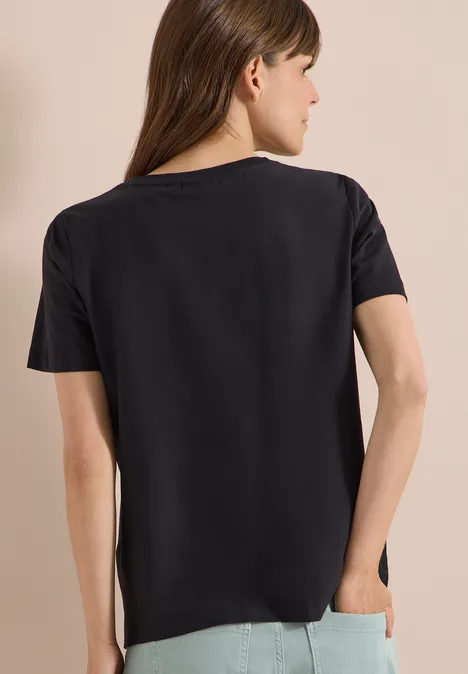 Frau in schwarzem Kurzarmshirt steht mit dem Rücken zur Kamera, der Hintergrund ist neutral.