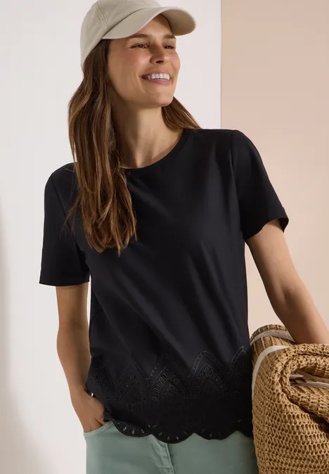 Frau mit schwarzem T-Shirt mit gewelltem Saum hält Strohtasche, vor hellem Hintergrund.