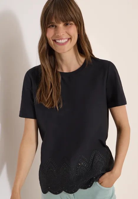 Eine Frau trägt ein schwarzes T-Shirt mit kurzen Ärmeln und gewelltem Lochmuster-Saum, Hand in Jeans-Tasche.