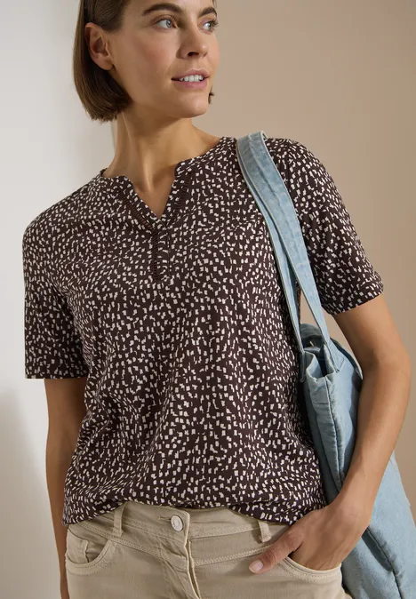 Frau in gemustertem dunkelblauem T-Shirt mit kurzen Ärmeln, heller Hose und blauer Schultertasche.