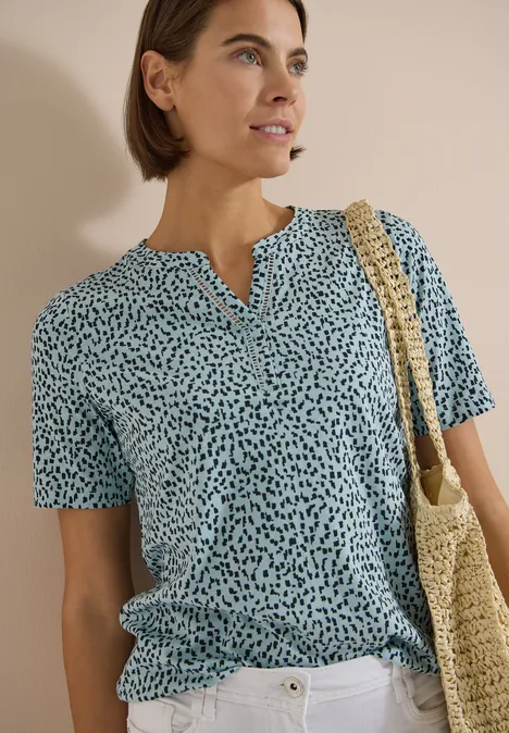 Frau im blauen, gepunkteten T-Shirt mit geflochtener Tasche vor neutralem Hintergrund.
