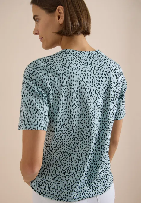 Rückansicht einer Person in locker sitzendem, hellblauem Shirt mit unregelmäßigen schwarzen Punkten.