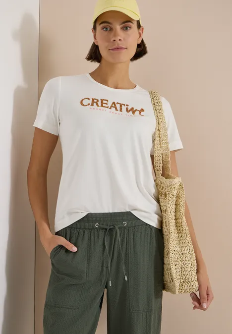 Person trägt weißes T-Shirt mit 'CREATIVE'-Aufdruck, grüne Hose und gehäkelte Tasche.