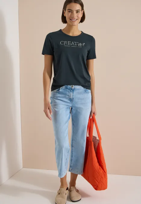 Frau in schwarzem T-Shirt mit Schriftzug, hellblauen Jeans und oranger Tragetasche vor neutralem Hintergrund.