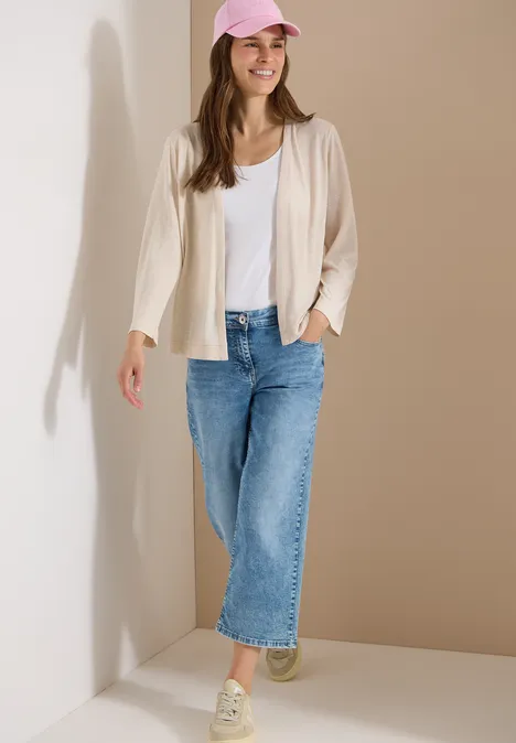 Veste ouverte en couleur unie avec manches 3/4 pearl beige Femme appuyée sur un mur clair, porte débardeur blanc, cardigan, jean bleu et baskets claires.