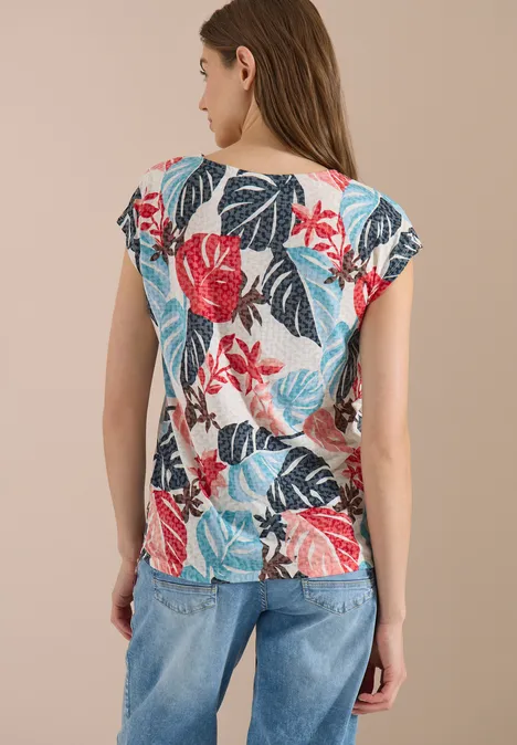 Frau von hinten trägt bunte, floral gemusterte Bluse mit kurzen Ärmeln und blaue Jeans vor neutralem Hintergrund.