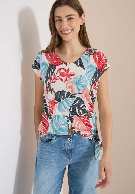 Frau in blauem Jeans und weißem T-Shirt mit buntem Blatt- und Blumenmuster vor hellem Hintergrund.