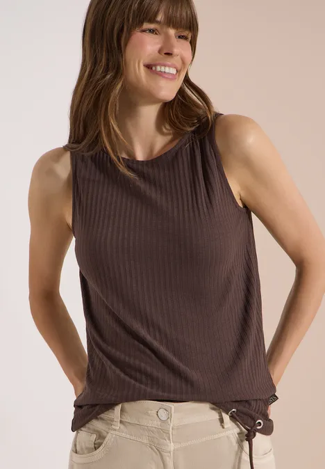Een vrouw draagt een bruin, mouwloos ribtopje en een lichte broek.