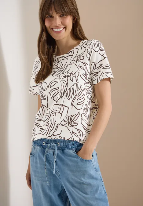 T-Shirt mit Rundhals und Blätterprint light alabaster white Frau in weißem T-Shirt mit Blattmuster und blauen, locker sitzenden Hose vor beigem Hintergrund.